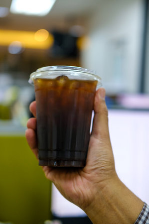 Close up hand holding a cup of ice black coffee or iced americano, cold brew coffee with blurred office background in the morningの写真素材