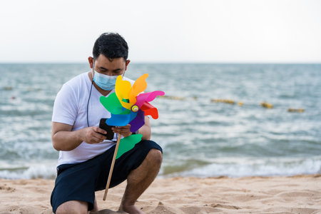 Rainbow pinwheel at beach and Asian man wear a mask sitting use smart phoneの写真素材