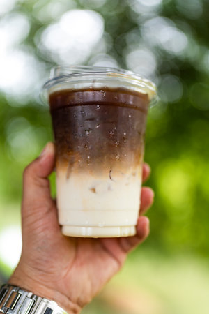 Hand holding the a plastic glass iced latte upper layer is brown bitter coffee and lower layer is fresh milkの写真素材