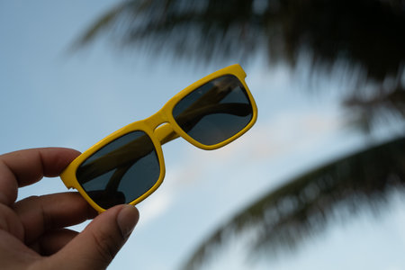 Hand holding yellow sunglasses held up to the sky at the beachの写真素材