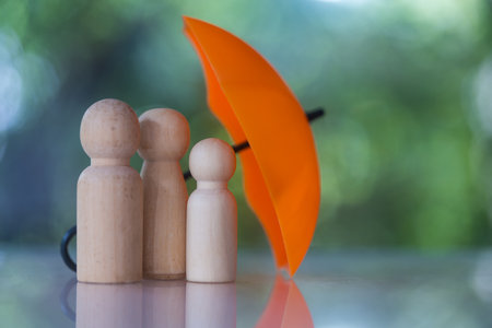 A family of wooden dolls are hiding under a orange umbrella, protecting wooden peg dolls.の写真素材