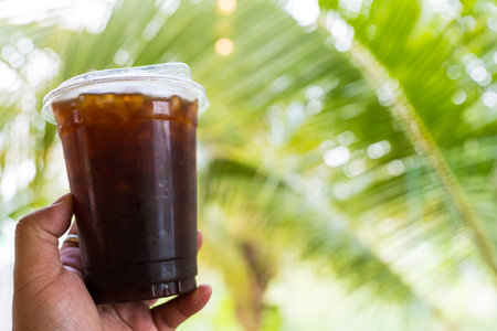 Close up of hand holding a plastic cup of black coffee on a nature blur backgroundの写真素材