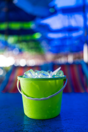 Full of ice cubes in green plastic bucket on blue wood table and blur of beach table and umbrellaの写真素材
