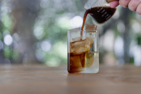 Hand pouring coffee into a glass tumbler with coffee with ice, on a wooden table with a nature background, the shot is taken from a frontal angleの写真素材