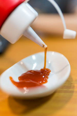 Person squeezing ketchup from bottle dispenser onto a pristine white ceramic plate.