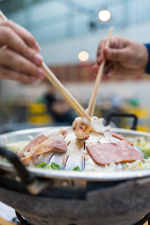 A hand expertly holding chopsticks, cooking meat over a steaming Moo Kata grill, where sizzling meats and bubbling broth create a unique Thai BBQの写真素材