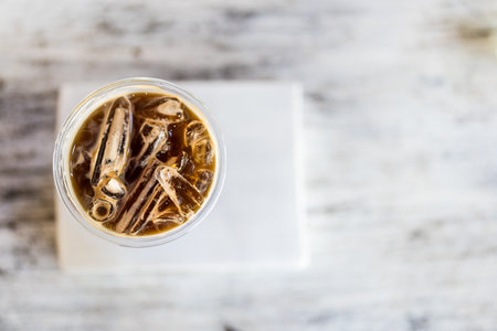A coffee cup with an iced Americano is placed on a minimal white wood background. Directly above...