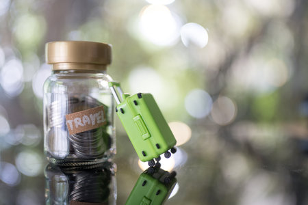 Travel label on glass with coins jar and green suitcase, highlighting budgeting and financial planning.の写真素材