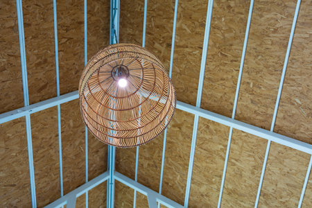 A light fixture made of woven material hangs from a ceiling. An intricately woven pendant lamp emits a warm, inviting glow, illuminating a space with its natural texture and unique design.の写真素材