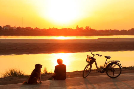 Silhouetted man with a dog watching sunset at Mekong river waterfront, Vientiane, Laosの写真素材