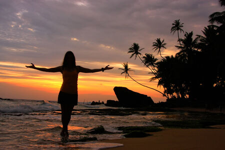 Silhouetted woman on a beach with palm trees and rocks at sunset, Unawatuna, Sri Lankaの写真素材