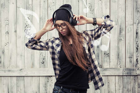 Hipster girl in glasses, headphones and black beanie on the wooden background