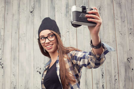 Hipster girl in glasses and black beanie making selfie with vintage camera on the wooden backgroundの写真素材