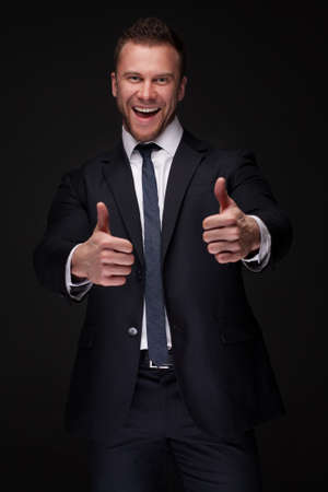 Portrait of young happy businessman with thumbs up isolated on dark backgroundの写真素材