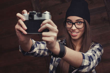 Hipster girl in glasses and black beanie with vintage camera making a selfie shot on the wooden の写真素材