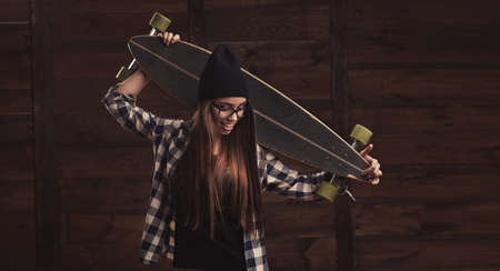 Hipster girl in glasses and black beanie with a longboard on the wooden backgroundの写真素材