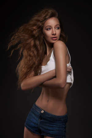 studio portrait of young woman with curly hair in white skirt and blue shorts on dark backgroundの写真素材