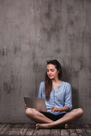 Young smiling woman sitting on the wooden floor with laptop near grunge grey wall. glasses on her headの写真素材