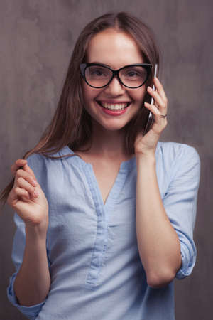 portrait of smiling woman with glasses speaking by cellphone near grey background wallの写真素材