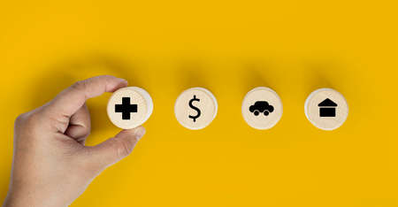 A person's hand holds a circular wooden block showing an insurance icon placed against a wooden block, money icon, car and house. Insurance concept. Clipping path and copy space.の写真素材