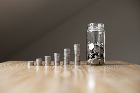 The glass jar is on the table, inside the jar there are many coins and there are piles of coins placed low to high on the side. Saving money for the future and using it in emergencies. Saving ideas.の写真素材