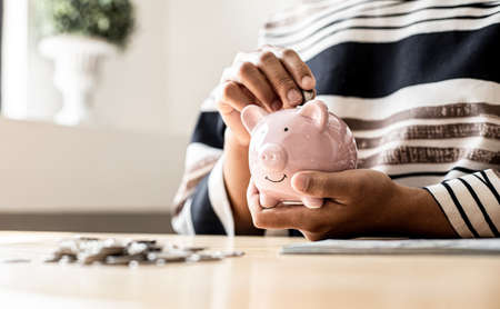 Woman putting a coin into a piggy bank in the shape of a pink pig. On her desk there are piles of coins that line from low to high, saving money for growth. Money saving ideas for investing in funds.の写真素材