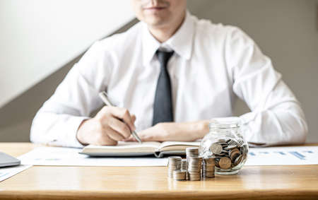 Piles of coins and a glass jar are placed on the desk of a man sitting in keeping a savings account. Placing coins in a row from low to high is a great saving for growth. Money-saving ideas.の写真素材