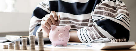 Woman putting a coin into a piggy bank in the shape of a pink pig. On her desk there are piles of coins that line from low to high, saving money for growth. Money saving ideas for investing in funds.の写真素材