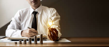 A businessman holds a light bulb and calculates his or her finances. He is making income and expenses and planning the use of the money. Calculate your energy consumption to save. Cost saving concept.の写真素材