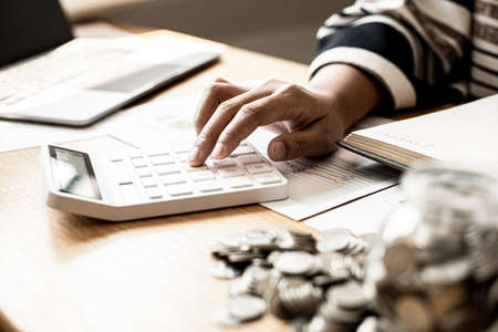 A woman is pressing a calculator to calculate expenses, she is doing an income-expense account and a money-saving plan to buy funds to make her money grow. Money saving ideas for investmentの写真素材
