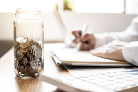 A glass jar with many coins on the desk of a businessman, financial planning by bringing the monthly savings into funds to make money grow faster. Concept of saving money for investment.の写真素材