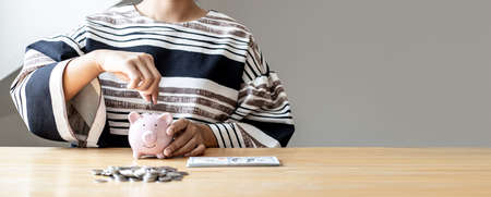 Woman putting a coin into a piggy bank in the shape of a pink pig. On her desk there are piles of coins that line from low to high, saving money for growth. Money saving ideas for investing in funds.の写真素材
