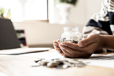 A woman is using two hands holding a glass jar with lots of coins inside, she has an income and expense accounting to plan her finances. Personal accounting ideas.の写真素材