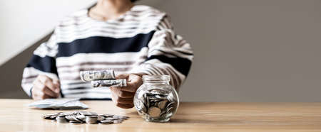 Woman holding dollar bills, on the table a large pile of coins and banks, she is managing to divide the money to save money and invest it to make it grow even more. Saving money and investing concept.の写真素材