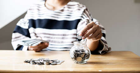 A woman is putting coins in a jar with a lot of coins inside, she is managing to divide her money to save money and invest it to make it grow even more. Concept of saving money and investing.の写真素材