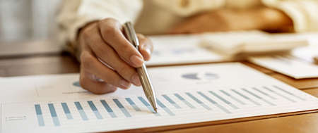Close-up businesswoman's hand holding a pen pointing at a bar chart on a corporate financial information sheet, the businesswoman examines the financial information provided by the finance department.の写真素材