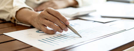 Close-up businesswoman's hand holding a pen pointing at a bar chart on a corporate financial information sheet, the businesswoman examines the financial information provided by the finance department.の写真素材