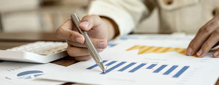Close-up businesswoman's hand holding a pen pointing at a bar chart on a corporate financial information sheet, the businesswoman examines the financial information provided by the finance department.の写真素材