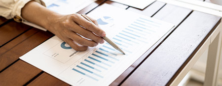 Close-up businesswoman's hand holding a pen pointing at a bar chart on a corporate financial information sheet, the businesswoman examines the financial information provided by the finance department.の写真素材
