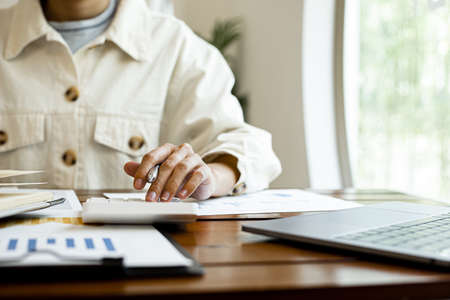 Close-up of a businesswoman using a white calculator, a financial businessman examining the numerical data on a company financial document, she uses a calculator to verify the accuracy of numbers.の写真素材