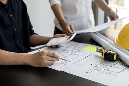 The architect was pressing the white calculator and there was an engineer standing there holding the floor plan. They are examining the draft blueprint for the designed building.の写真素材