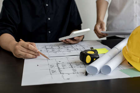 The architect was pressing the white calculator and there was an engineer standing there holding the floor plan. They are examining the draft blueprint for the designed building.の写真素材