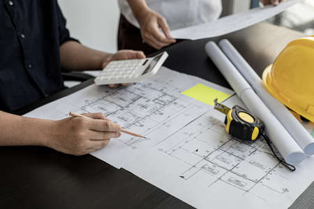 The architect was pressing the white calculator and there was an engineer standing there holding the floor plan. They are examining the draft blueprint for the designed building.の写真素材