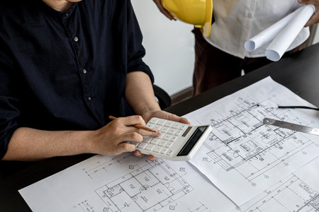 The architect was pressing the white calculator and there was an engineer standing there holding the floor plan. They are examining the draft blueprint for the designed building. Construction and interior ideas.の写真素材