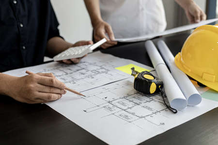 The architect was pressing the white calculator and there was an engineer standing there holding the floor plan. They are examining the draft blueprint for the designed building.の写真素材