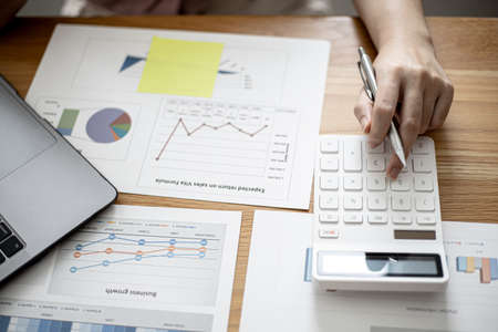Businesswoman is using a calculator to check the numbers on a company financial document during a meeting with shareholders, corporate finance documents showing a bar graph format. Financial concept.の写真素材