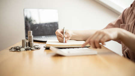 A woman is writing her income and expenses in her book, she keeps a monthly account to plan her...
