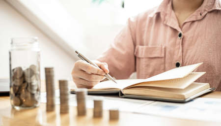 A woman is writing her income and expenses in her book, she keeps a monthly account to plan her savings and invest the money. Concept of saving money and investing it to grow money.の写真素材