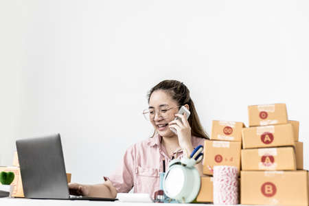 A woman is talking on the phone and responding to a customer chat through the company's online sales page, she is taking orders and preparing packs for delivery by shipping company.の写真素材