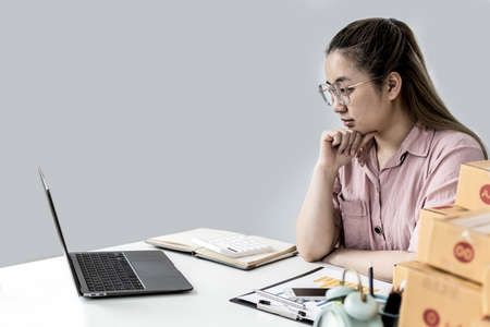 Online shop owners are sitting, checking the order information from her page, she is viewing information from the laptop on the table with a large number of parcel boxes. Product sales concepts onlineの写真素材